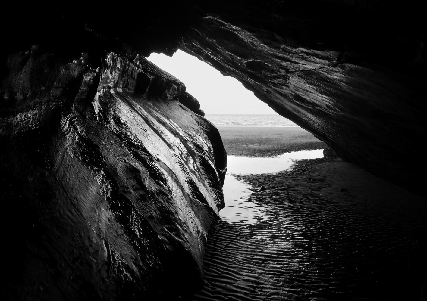 Black and white photograph by Mark Robert Davey showing a cave from the inside looking out towards the beach and sea.