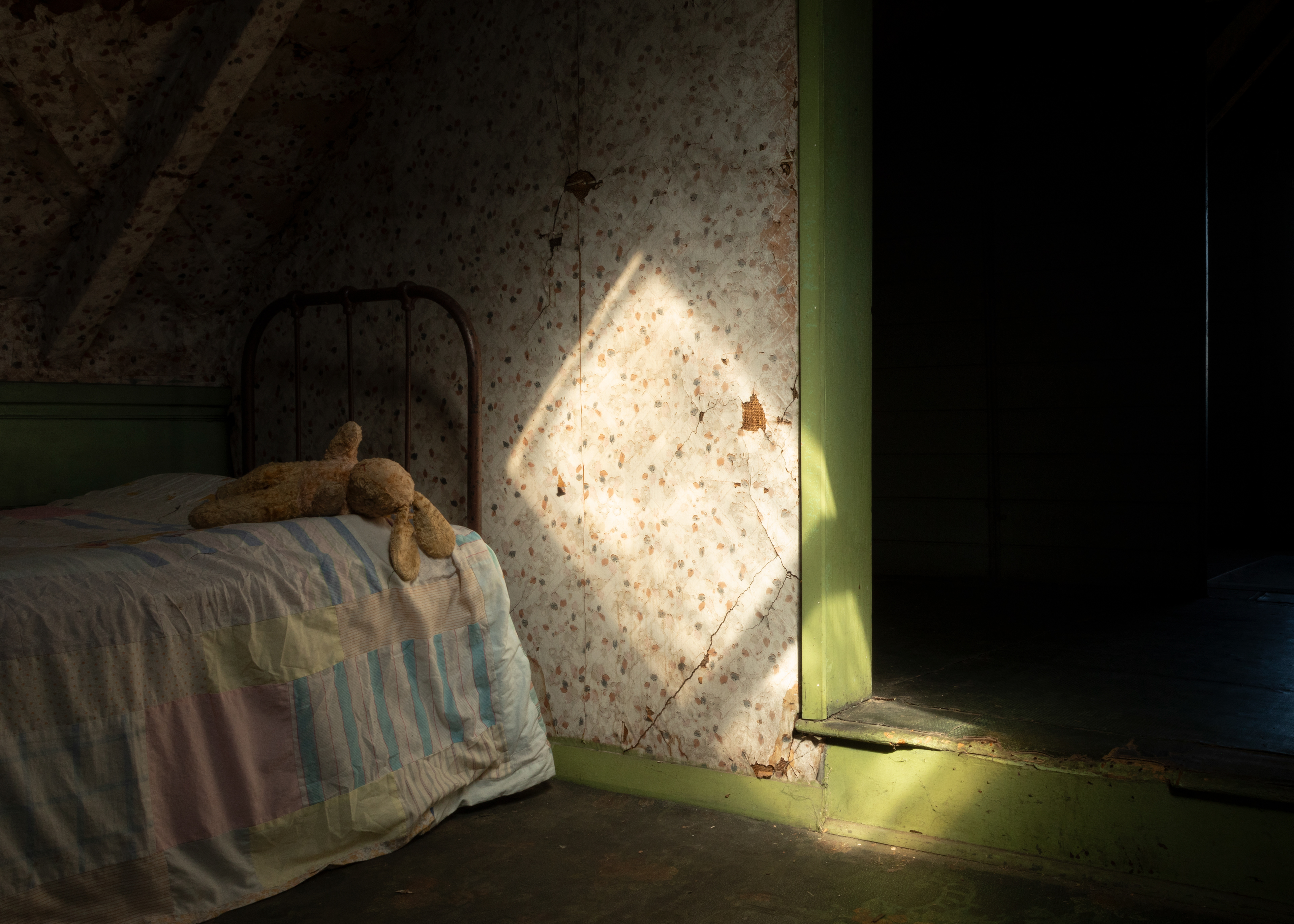 Interior of historic bedroom with green skirting. Worn linoleum floor and vintage wallpaper. A rectangle of light hits the wall pointing the viewer towards a vintage bed, dressed with a pale quilt and worn vintage rabbit soft toy 