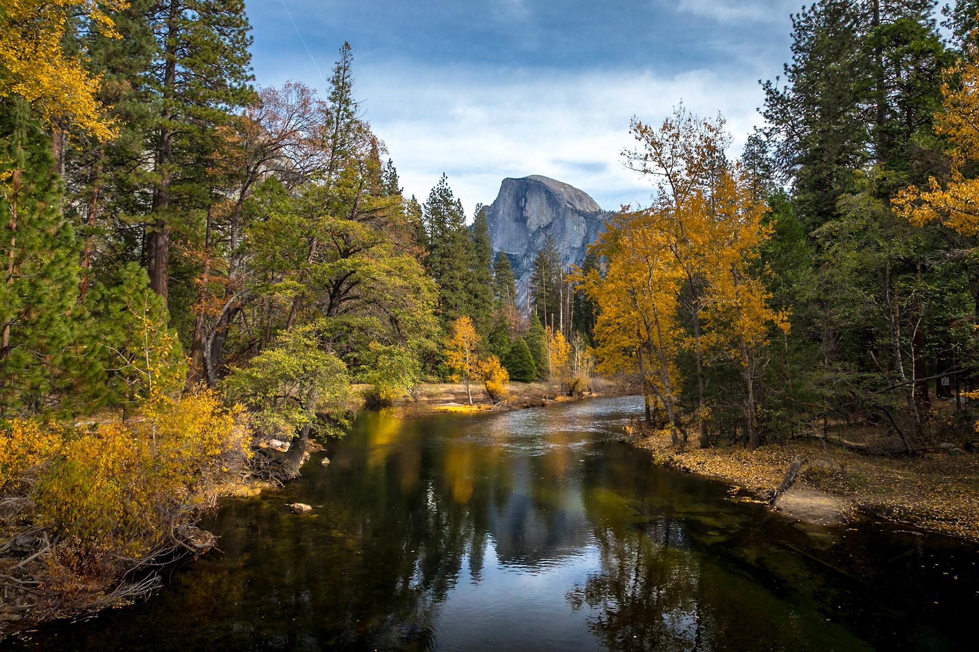 A tranquil autumn view captures Half Dome reflected in the peaceful waters of Yosemite Valley, surrounded by vibrant foliage and towering trees.