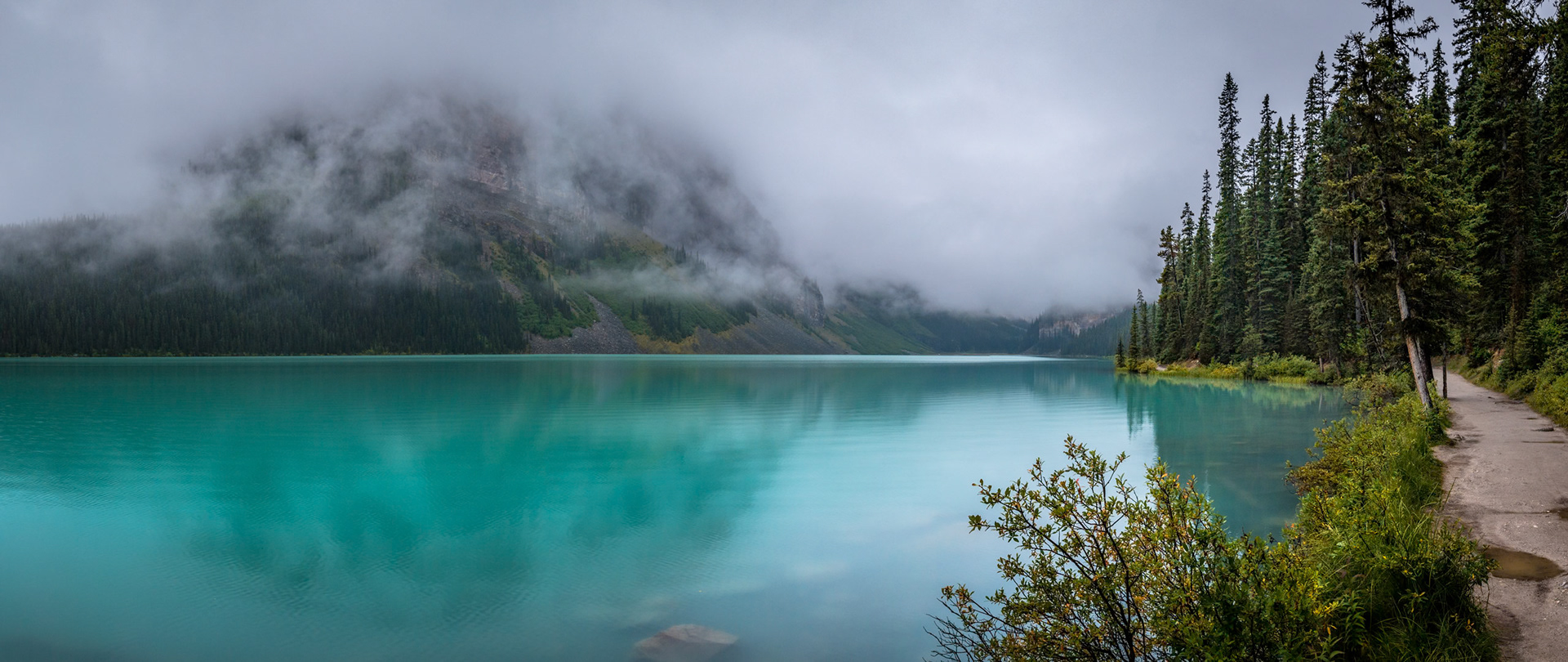 Scenic view of Lake Louise and Fairview Mountain with trees surrounding the lakeshore on a foggy day, Banff National Park, Alberta, Canada