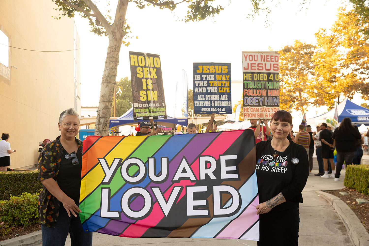 Janie Acosta (she/her), left, and Jana Zimmelman (she/her), right, are both part of the Long Beach Christian Fellowship. The two hold up a ‘You are loved’ sign in front of protestors at the Downey Pride Festival on August 10th, 2024. Recognizing the damage that the church has done to people in the LGBTQIA+ community, they want their sign to offer a message of hope.