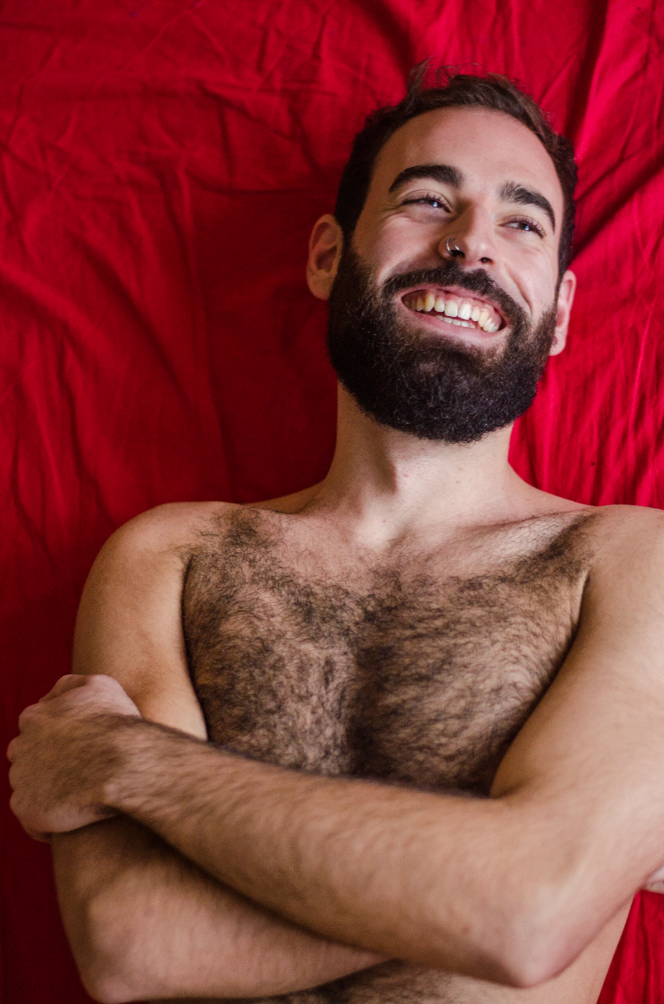 Bearded man on bed smiles. Male nudity, nude, man, portrait, chest, piercing, hairy, beard, naked. Nudez masculina, homem, pelado, barba, peludo, retrato, nu, peito, ri, sorriso.