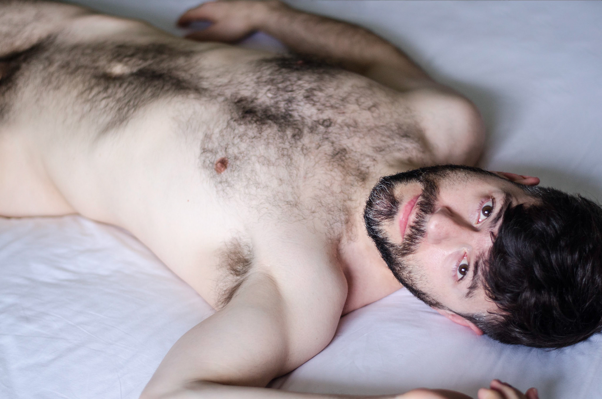 Naked hairy man lies on bed, stares at the camera. Portrait, nude, photography, retrato, fotografia, nu, homen, barba, penis, grisalho