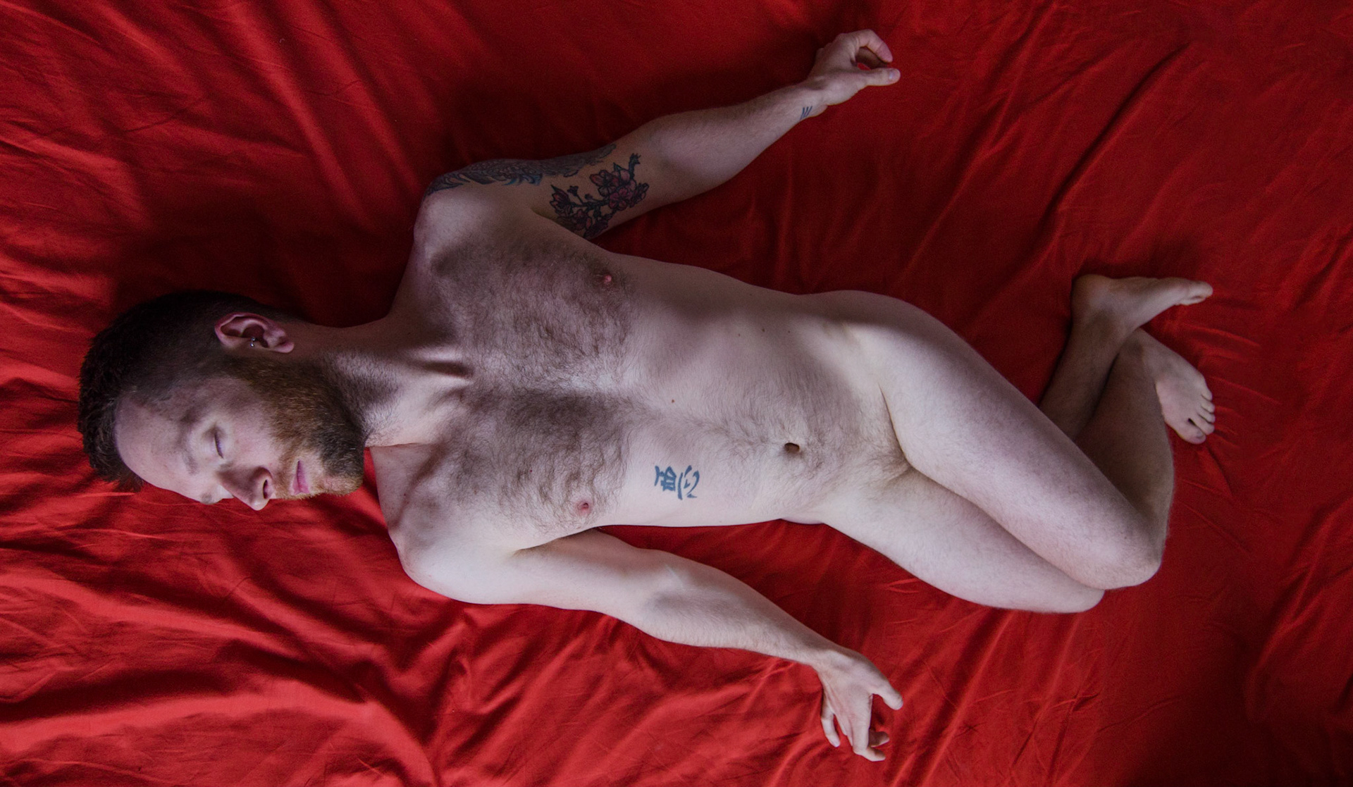 Undressed ginger man, crossed legs, lies on bed. Male nudity, nude, man, portrait, hairy, beard, naked. Nudez masculina, homem, pelado, barba, peludo, retrato, nu. Pernas cruzadas, cama vermelha
