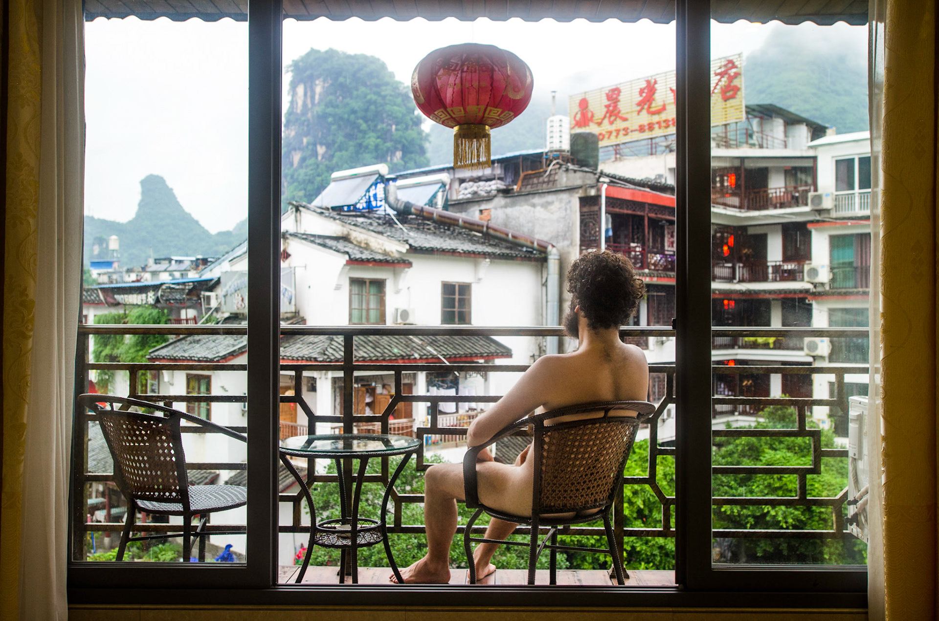 Em uma varanda, figura masculina de barba admira a vista em algum lugar no interior da China. On a balcony, a male figure with a beard observes the landscape somewhere in China countryside. Projeto contínuo de auto retrato. China, transição, viagem, montanhas, lanterna, nu, pelado, pelos, cabelo, cabeludo Ongoing self portrait project. transition, mountain, lantern, naked, nude, hair, hairy, body hair