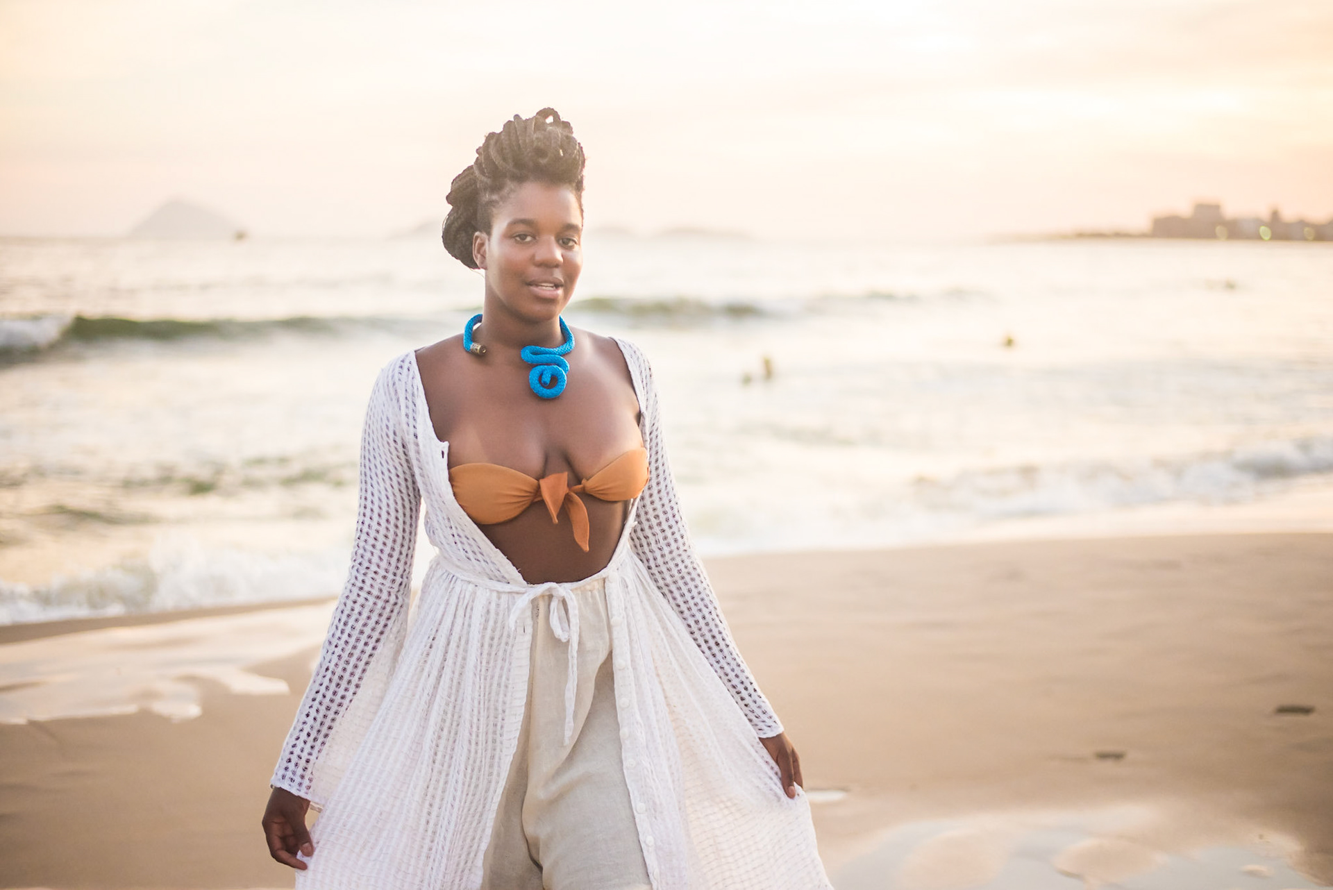 Model with orange top, blue necklace, white pants and dress poses to camera. mulher de laranja, colar azul, vestido e calça branca posa para foto. fashion photography, designer, clothes, dress, accessory, product, still, brand, lookbook, shooting, vibrant, colorful, beach, creative, catalogue, outfit, look.fotografia de moda, design, roupa, vestido, acessório, produto, still, marca, vibrante, colorida, praia, catálogo, rio de janeiro.