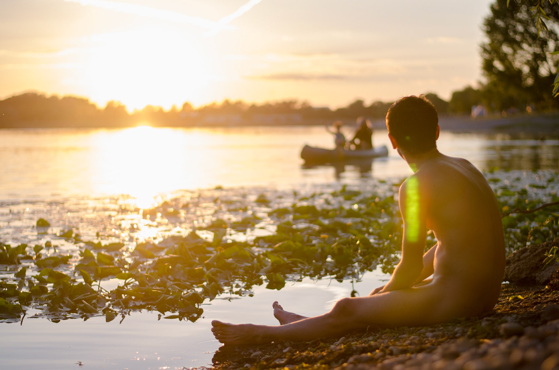 Naked man watches sunset from beach. Homen nu assiste por-do-sol de praia. Male nudity, nude, man, portrait, hairy, beard, naked, row, boat. Nudez masculina, pelado, barba, peludo, retrato, barco a remo.