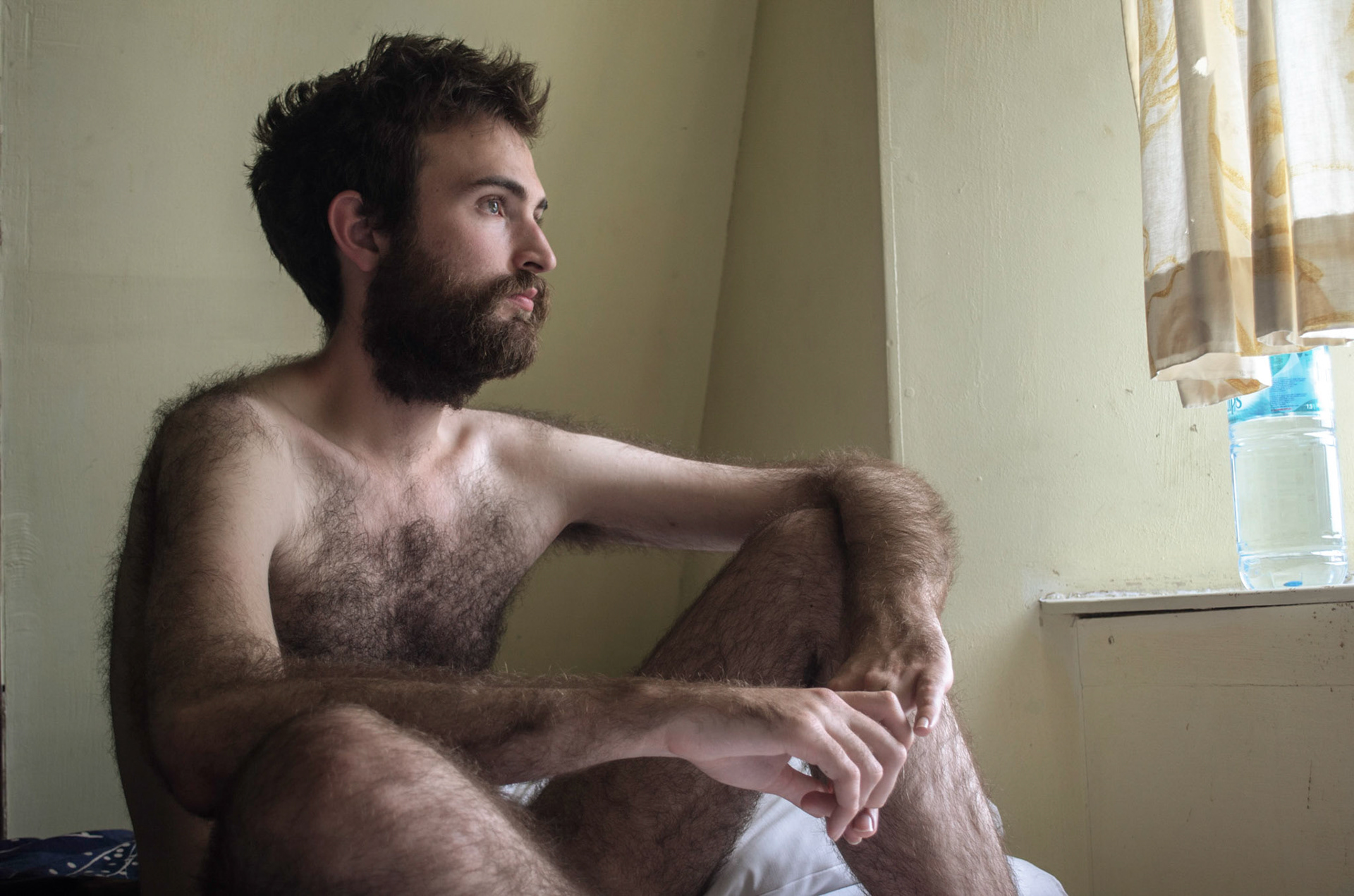 Naked hairy man sits on hotel bed and stares at window. Portrait, beard, male. hair. Homen nu peludo olha janela de hotel. retrato, nu, maculino, chico morais
