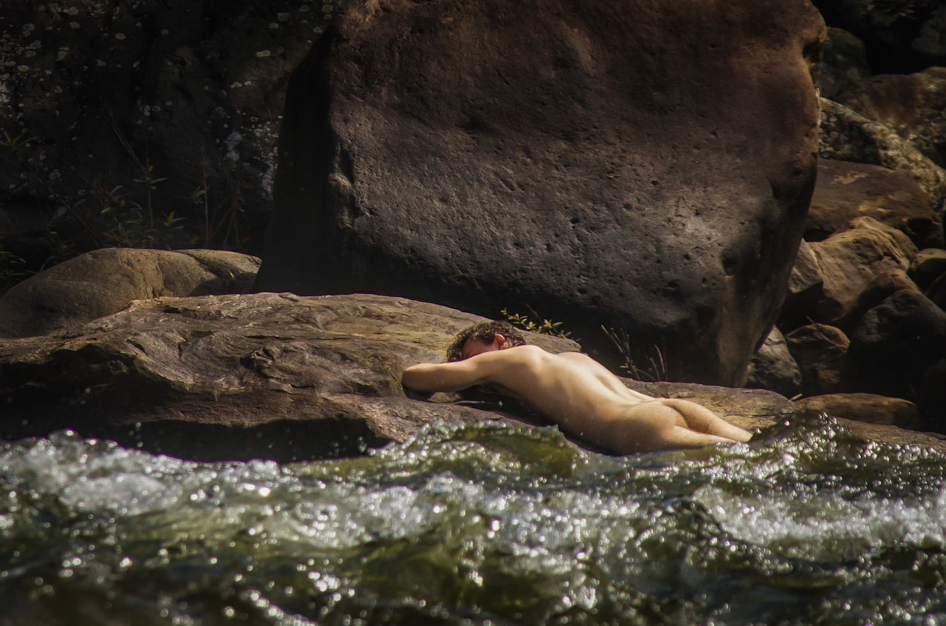 Man lies naked by giant river. Homem se deita nú na beira de grande rio. Male nudity, nude, man, portrait, arse, ass, naked. Nudez masculina, homem, pelado, bunda, nádegas, retrato, nu.