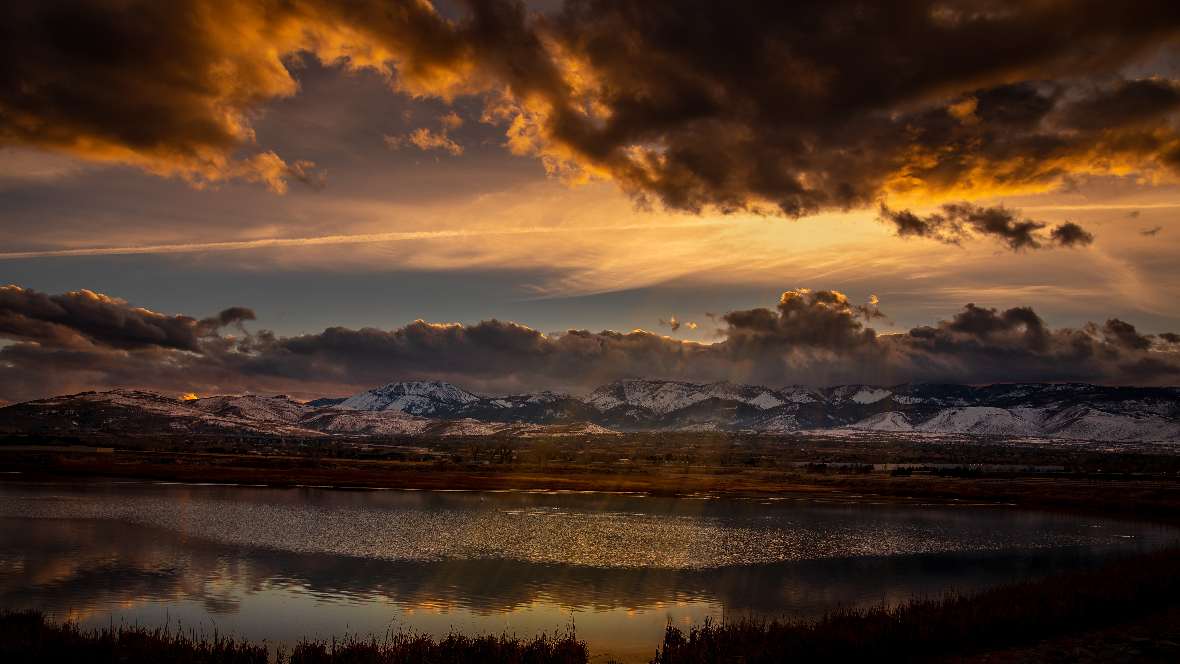 Nightfall approaches with a pond in the foreground, clouds over the mountains and looming immediately overhead, and a ray of sunlight breaking through the clouds.