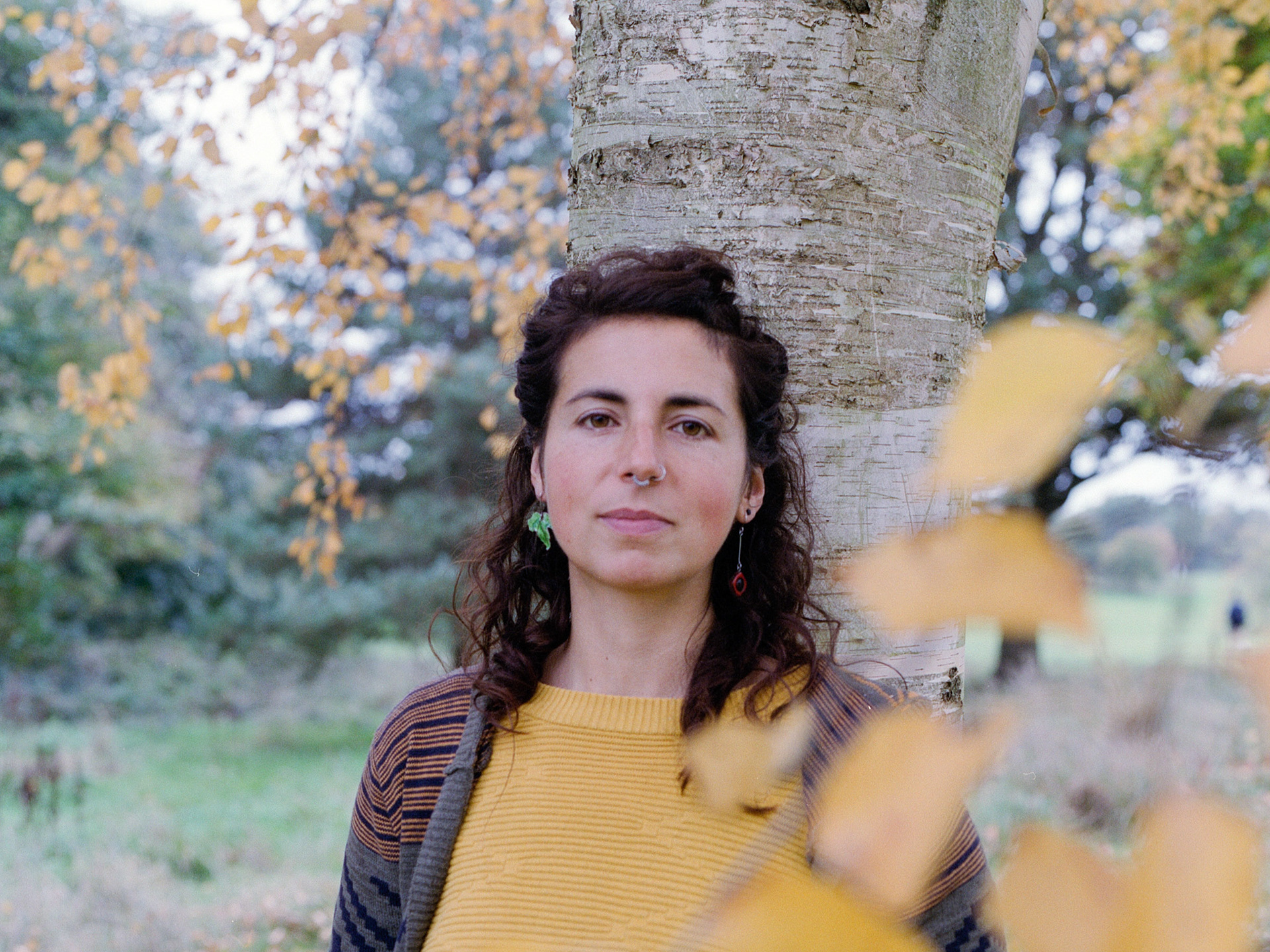 Ángela is leaning against a silver birch tree and looking into the camera. Her top is the same colour as the tree's leaves which have turned orange for autumn.