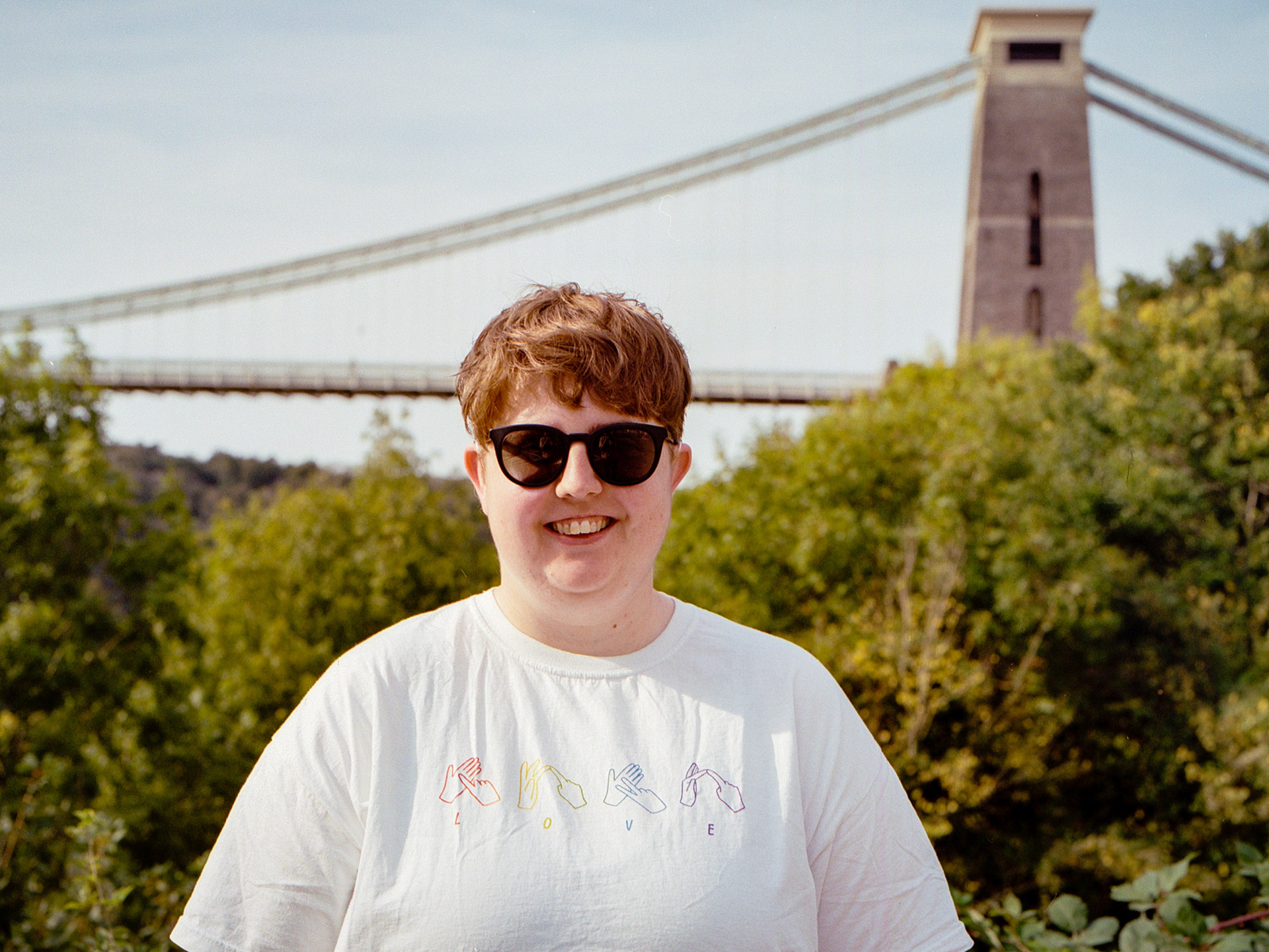 Charlotte is standing in front of Bristol's suspension bridge. She is wearing a t-shift that says LOVE on it. The letters to love have sign language above each letter.