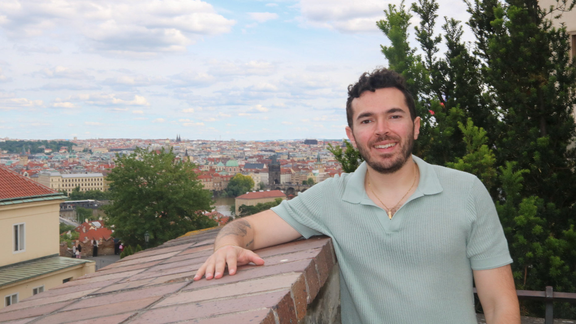 Zach Reider smiling and leaning on a stone wall overlook, with a panoramic view of Prague's red-roofed cityscape and the Vltava River in the background