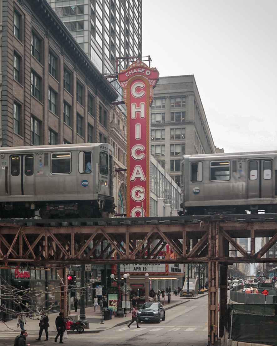 Chicago Theatre marquee on State Street with two CTA elevated trains passing in front, pedestrians below on an overcast day.Chicago Theatre marquee on State Street with two CTA elevated trains passing in front, pedestrians below on an overcast day