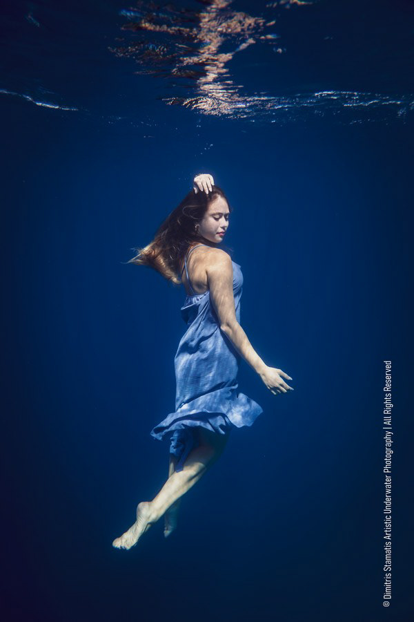 A young girl in a light blue dress floating underwater with one arm raised.
