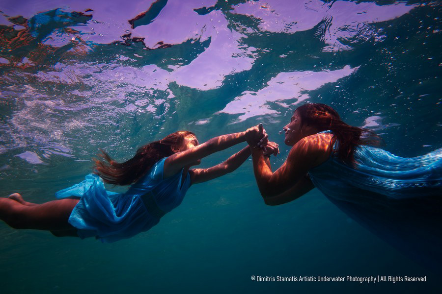 Mother and daughter in blue dresses underwater holding hands.