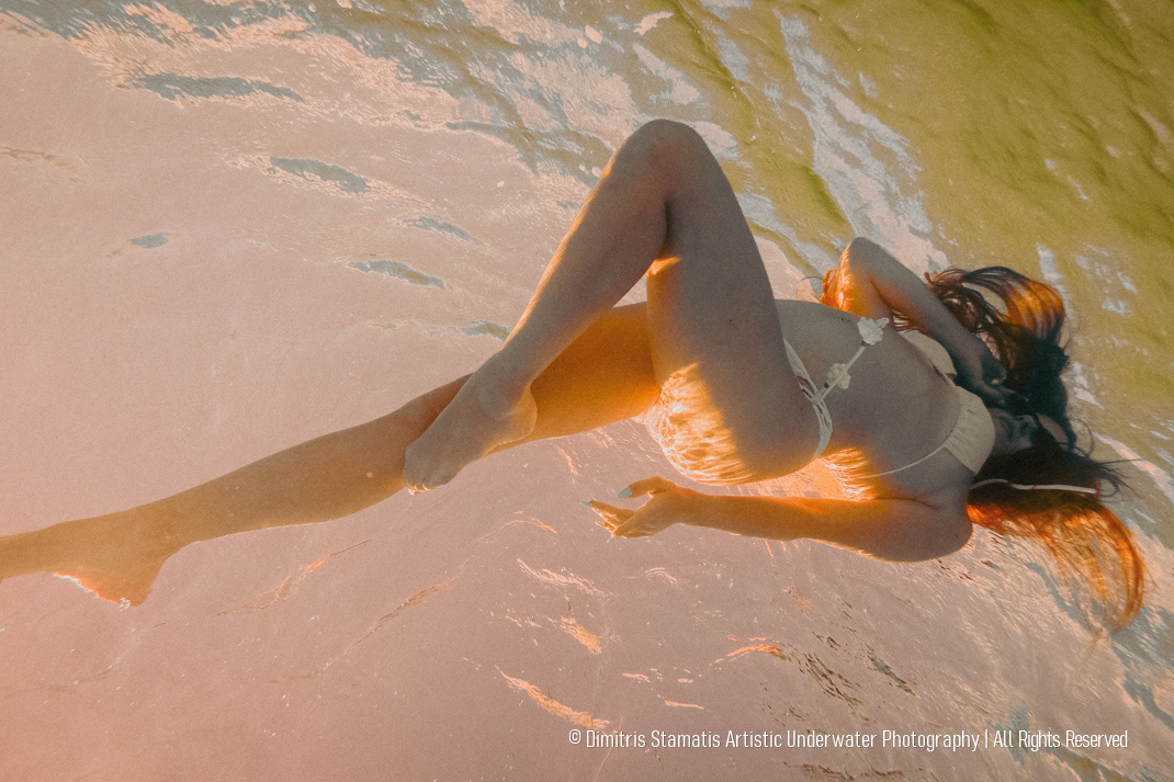 A young girl wearing a light-colored swimsuit floats gracefully underwater beneath warm, golden light, with ripples on the surface creating soft distortions and an ethereal atmosphere.
