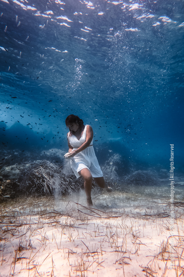 A woman wearing a white dress kneels on the sandy seabed underwater, moving the sand with their hands as particles drift upward among aquatic plants and small fish in clear blue light.