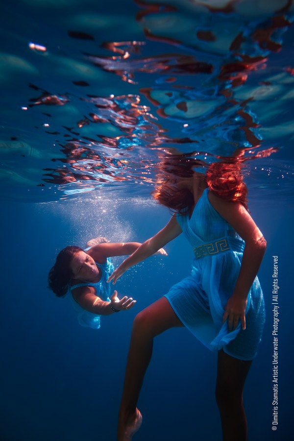 Mother and daughter in blue dresses underwater reaching toward each other.