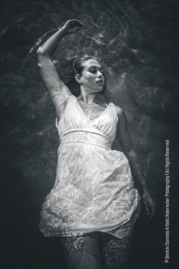 A young woman wearing a light, thin dress floats underwater while reflections and ripples of light move across the fabric and her body.