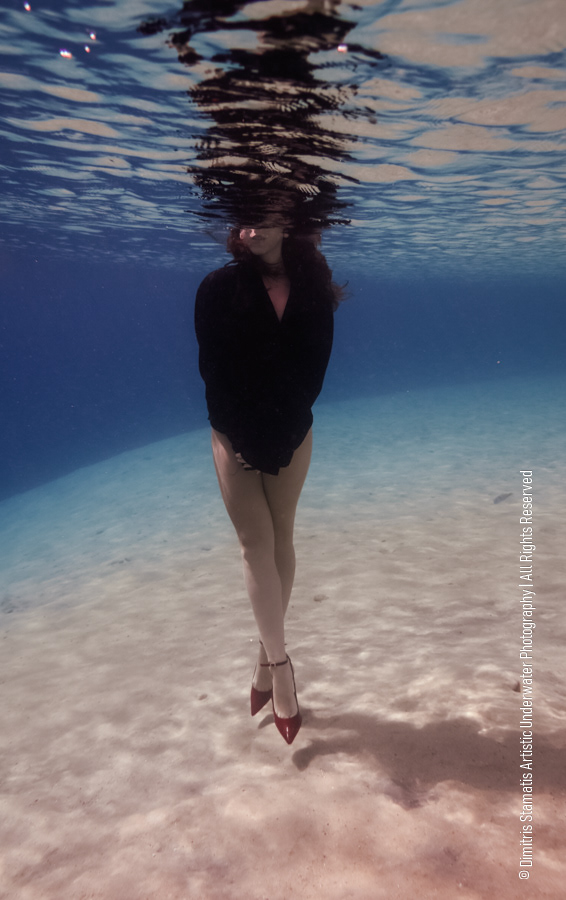 A girl underwater wearing a dark long‑sleeved top and red high‑heeled shoes, with arms crossed. 