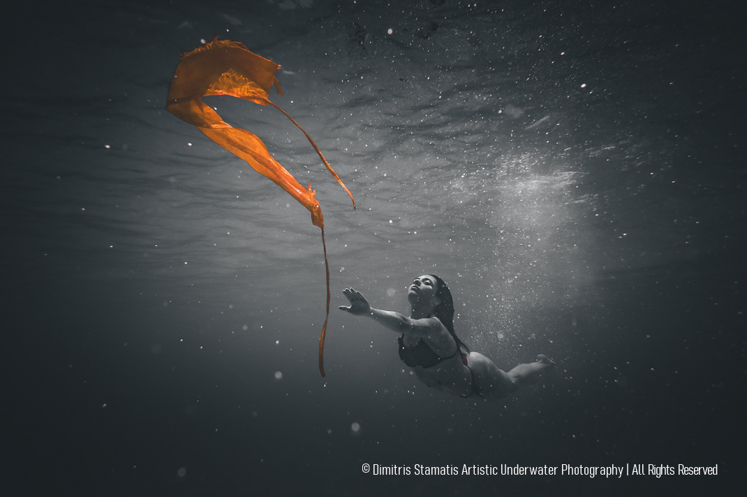 A young girl swims underwater toward a vivid piece of orange fabric, with the dark water creating strong contrast as light highlights the movement and texture of both the fabric and the surrounding water.