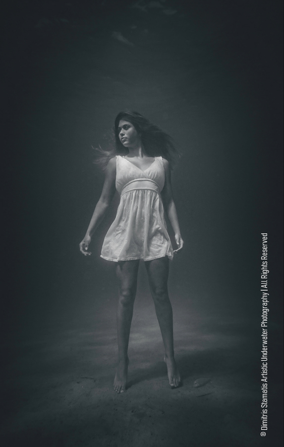 A young girl standing underwater on the sandy seabed in a light dress as soft diffused light and drifting hair create a calm, ethereal scene.