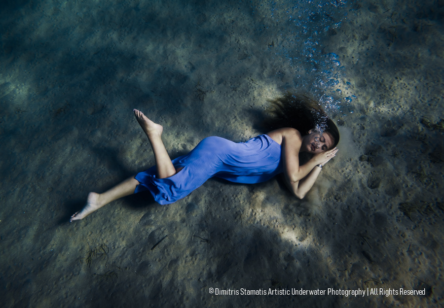 A woman wearing a blue dress lies on the sandy sea floor in a sleep‑like pose while small bubbles rise toward the surface.