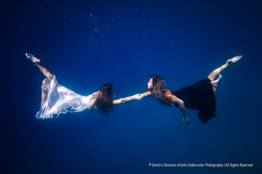 Two young girls underwater in flowing black and white dresses reaching out to hold hands.