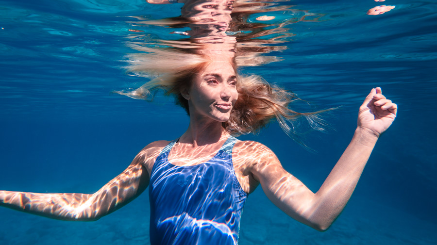 Şahika Ercümen underwater with flowing hair and sunlit patterns across her skin and blue swimsuit.