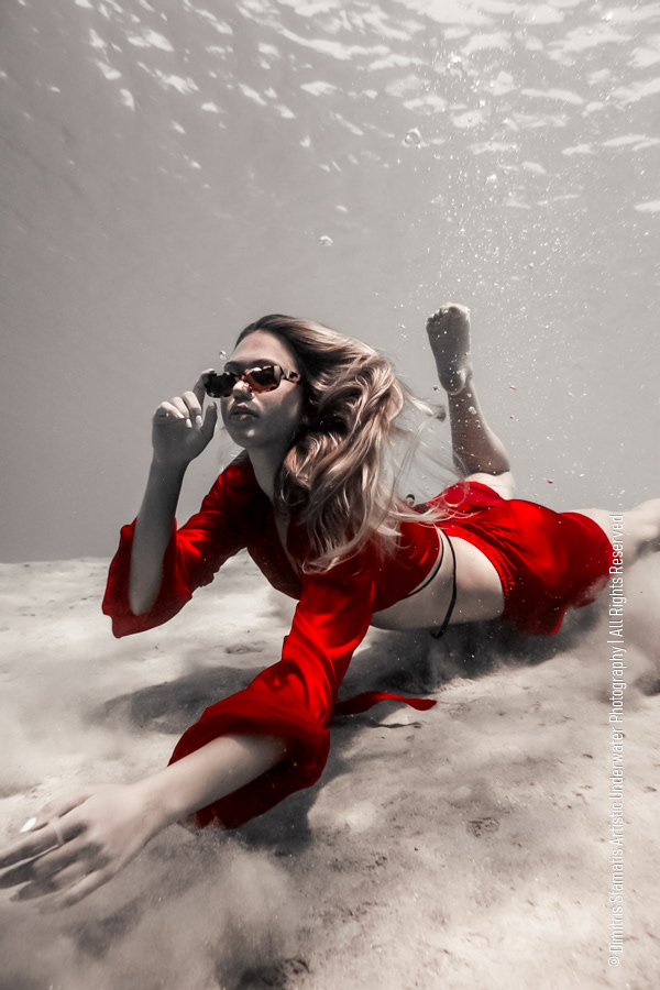 A young girl in a bright red and sunglasses outfit floating underwater above the sandy seabed as hair drifts and soft light filters through the water.