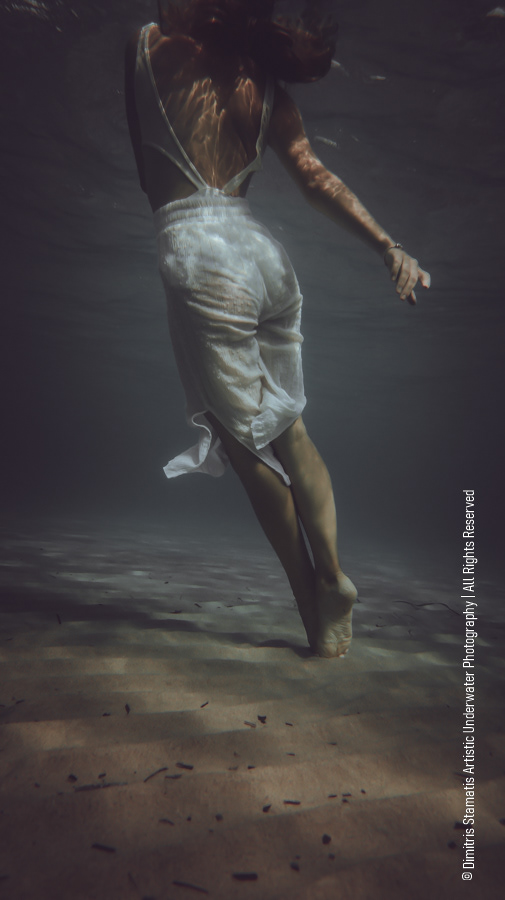 A young gir; wearing a light white dress floats upward underwater, viewed from behind, with legs extended and sunlight creating shimmering patterns on the skin and clothing above the sandy seabed.