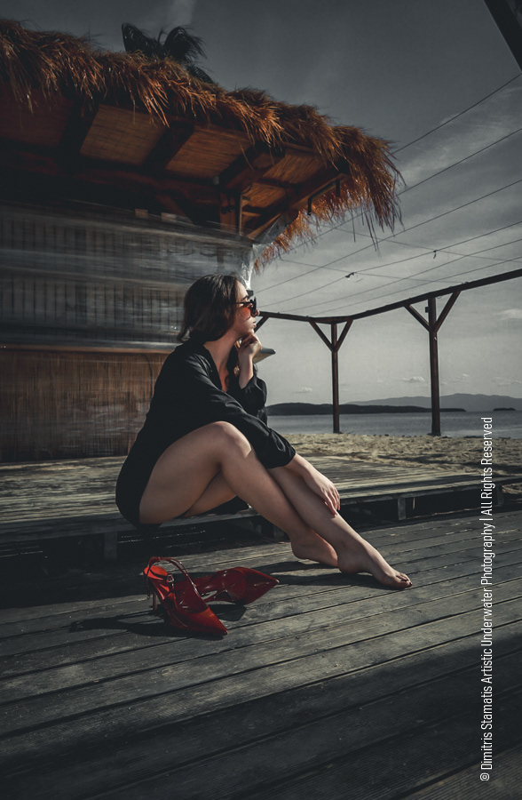 A young girl sitting barefoot on a wooden deck near a beach hut with red heels beside them.