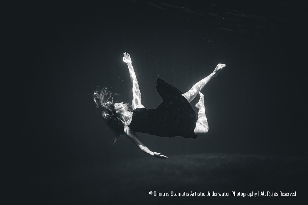 A young girl wearing a dark dress floats underwater with extended limbs, while shimmering light from above creates textured patterns on the skin and fabric against the dark background.