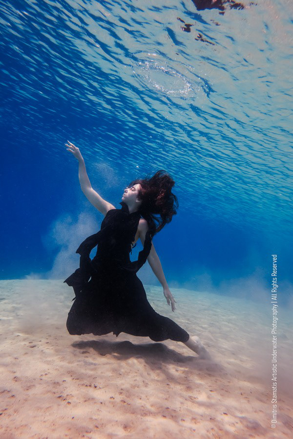 A girl underwater in a long dark dress kneeling on the sandy seafloor and reaching upward.