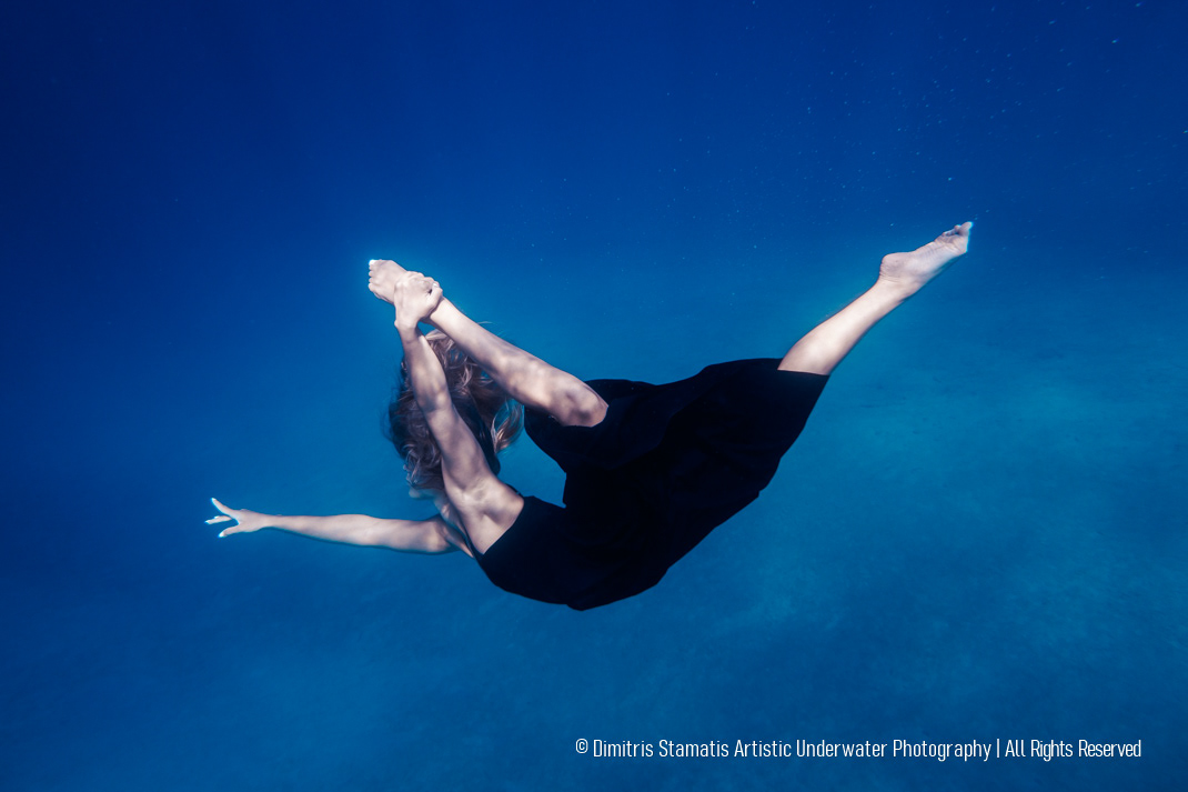 Young girl underwater in a black dress performs a ballet pose while holding her leg. 