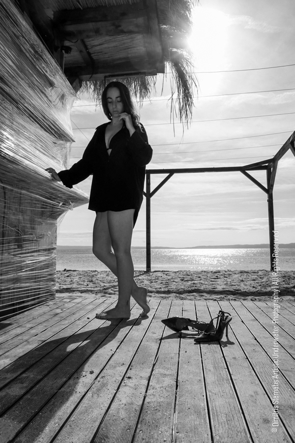 A young girl stands barefoot on a wooden deck by the beach wearing a dark long-sleeved shirt.