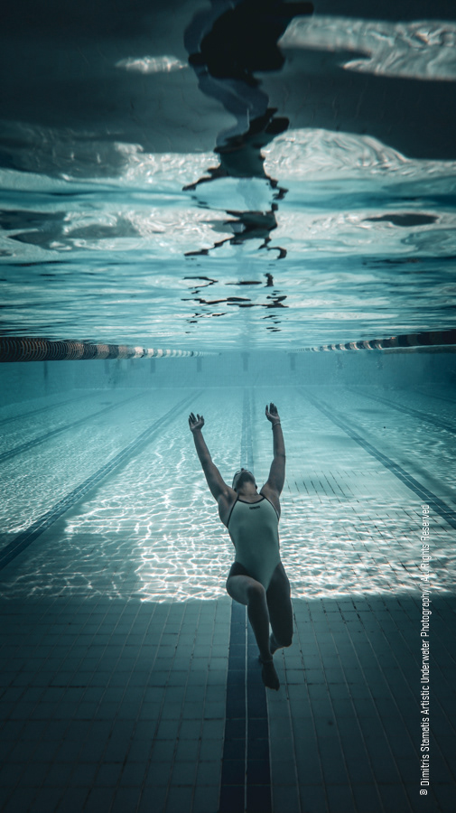 A swimmer wearing a light-colored swimsuit reaches upward underwater in a vertical position above the tiled pool floor as light patterns reflect across the scene.