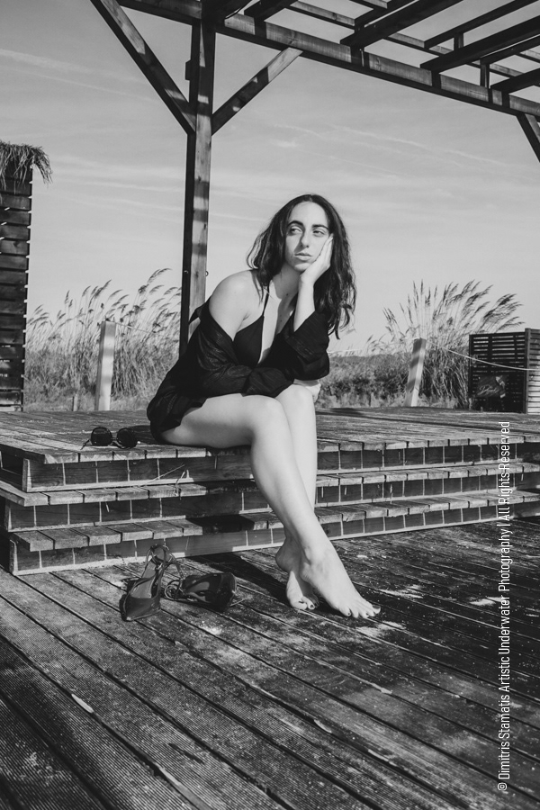 A girl in a dark outfit sitting barefoot on a brick structure outdoors beneath a wooden frame.