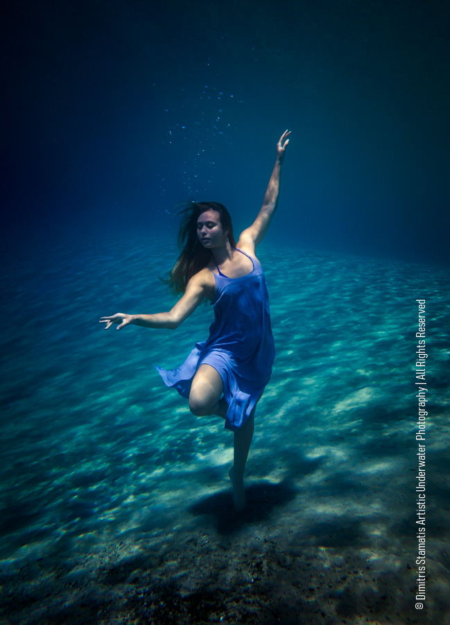A young girl underwater in a flowing blue dress moving as if dancing above a sandy seabed.