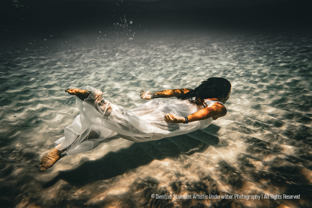 A young woman wearing a long, flowing white dress swims horizontally just above the sandy seabed underwater, with sunlight filtering through the surface and creating shimmering patterns on the sand and fabric.