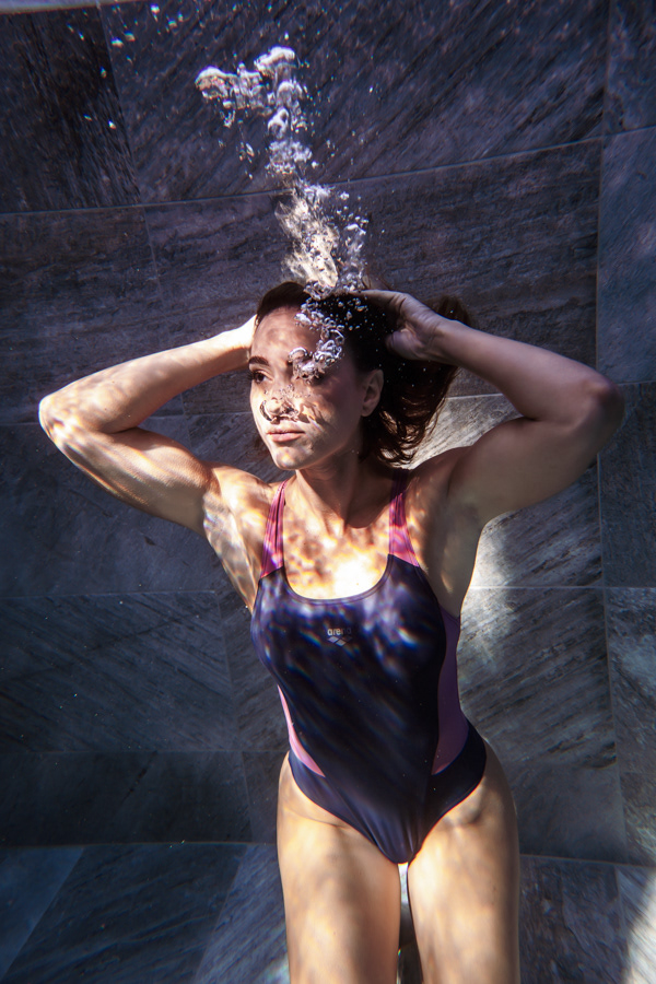 Maria Kirykou underwater in a pool with both hands on her head as bubbles rise toward the surface.
