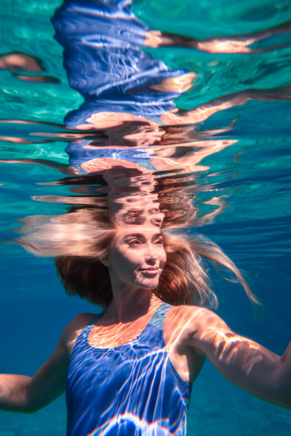 Şahika Ercümen underwater with her face mirrored by the rippling water surface, illuminated by sunlit reflections.