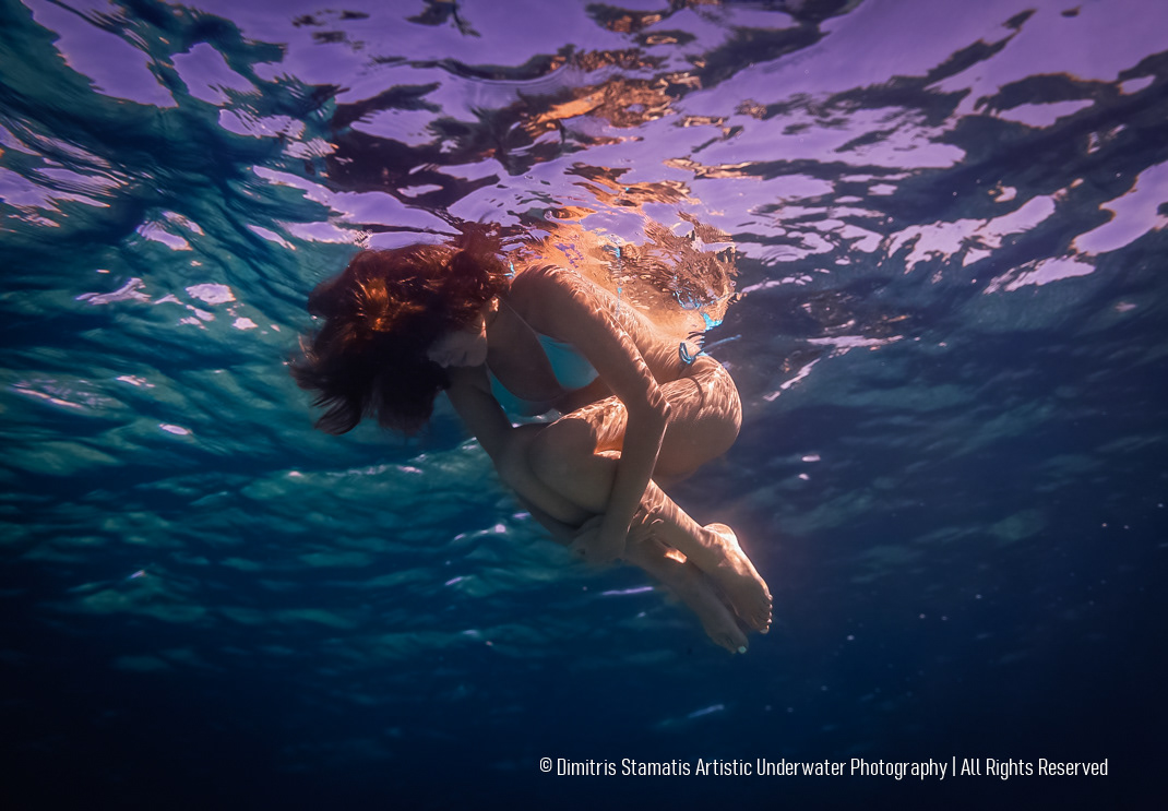 A woman wearing a turquoise swimsuit floats underwater in a curled position while colorful reflections of purple, blue, and gold light move across the surface and their body.