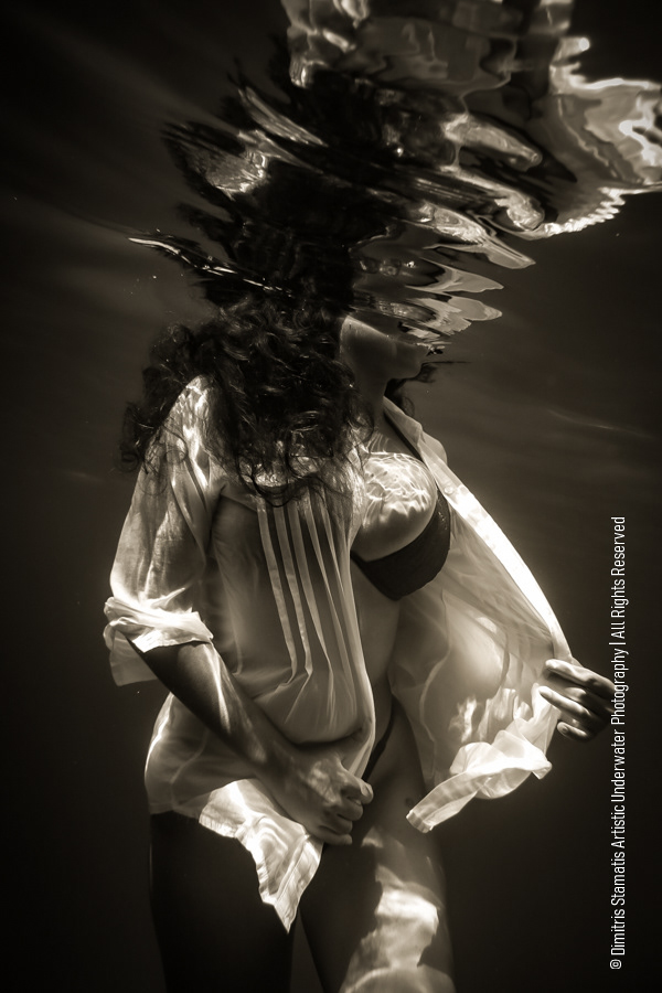 A young girl underwater in a light, semi‑transparent shirt with hair floating freely, while the surface above creates a rippled, distorted reflection in the clear water.