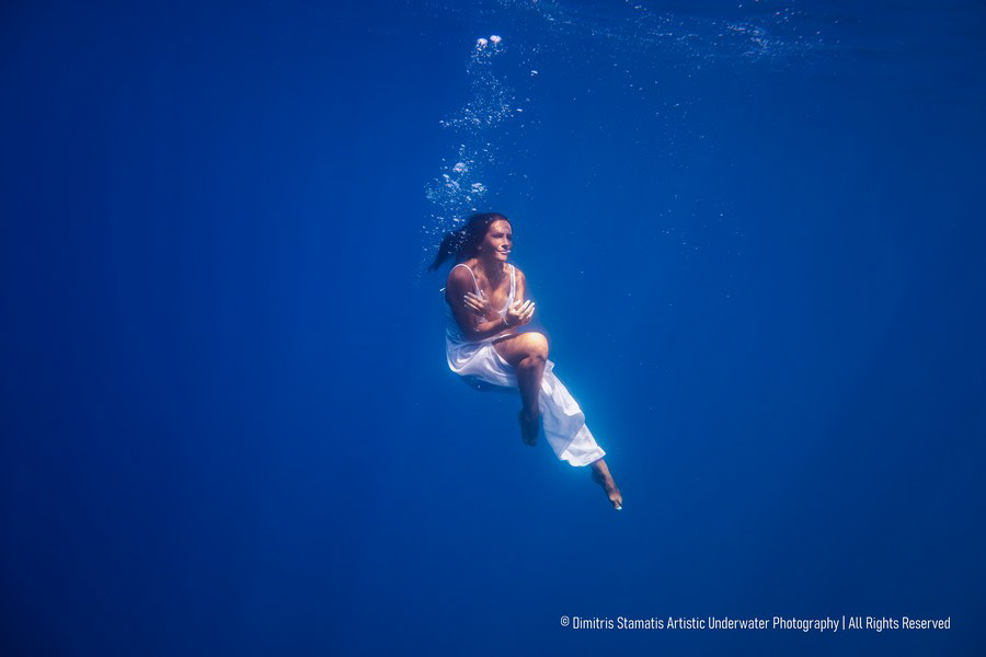 A young woman wearing a white dress suspended underwater in a deep blue sea.