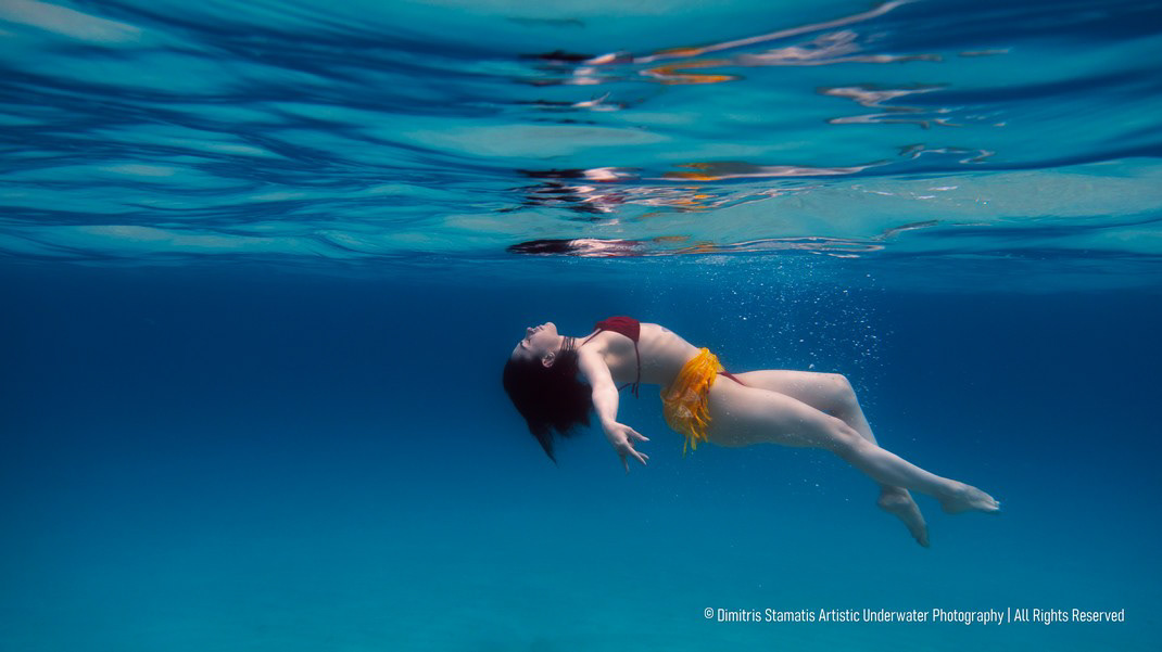 A woman in a red bikini top and yellow flowing scarf drifting underwater beneath the rippled surface, illuminated by soft blue light.