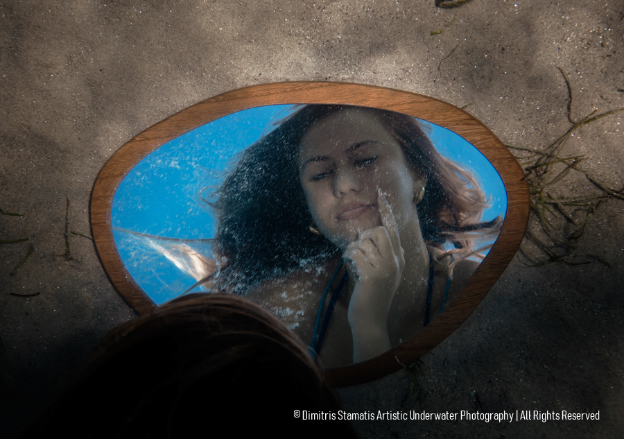 A round mirror resting on underwater sand reflecting a girl's face with one hand touching her chin.