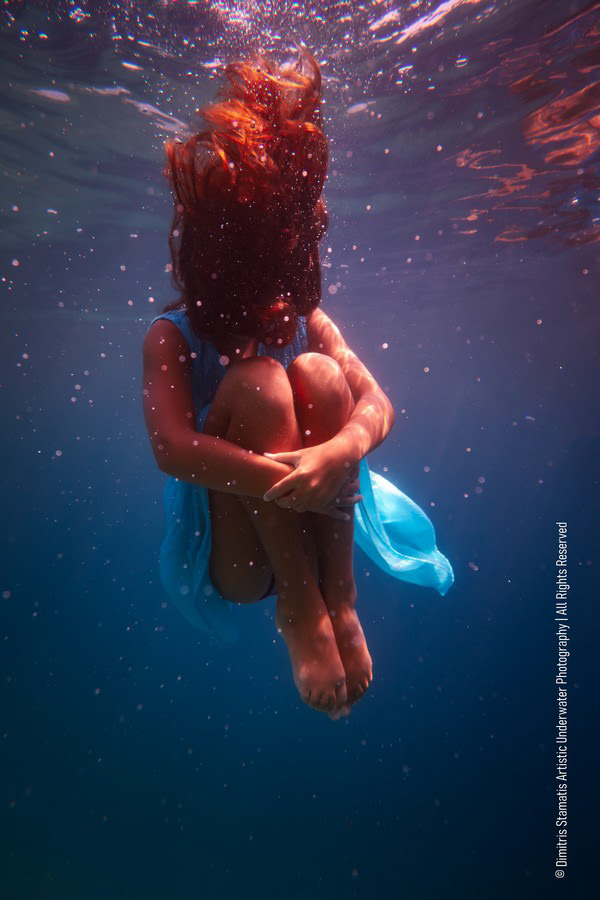 A young curled underwater with floating red hair and a light blue dress.