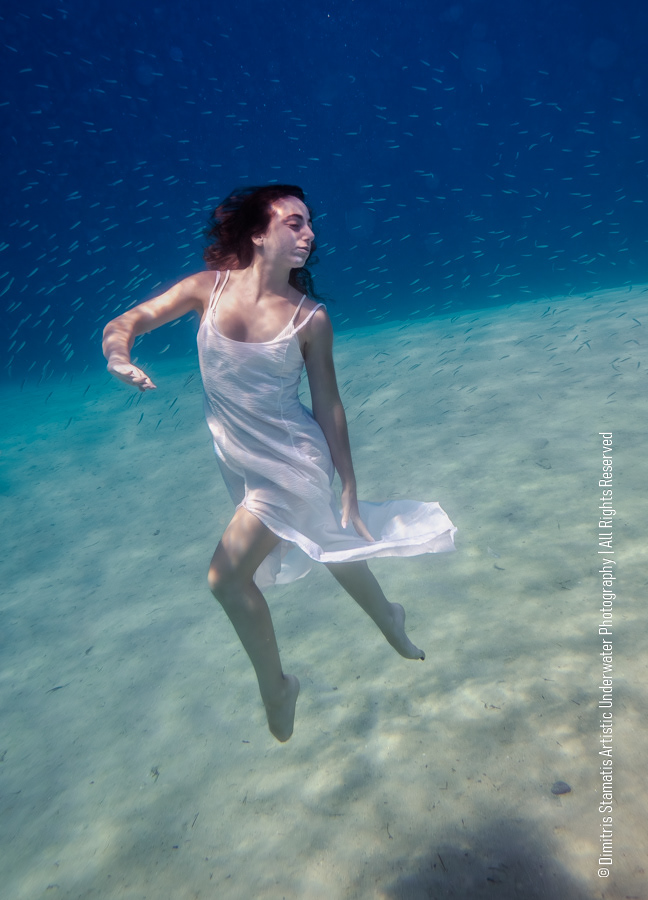 A girl underwater wearing a white dress floats gracefully above a sandy seabed, surrounded by fish.